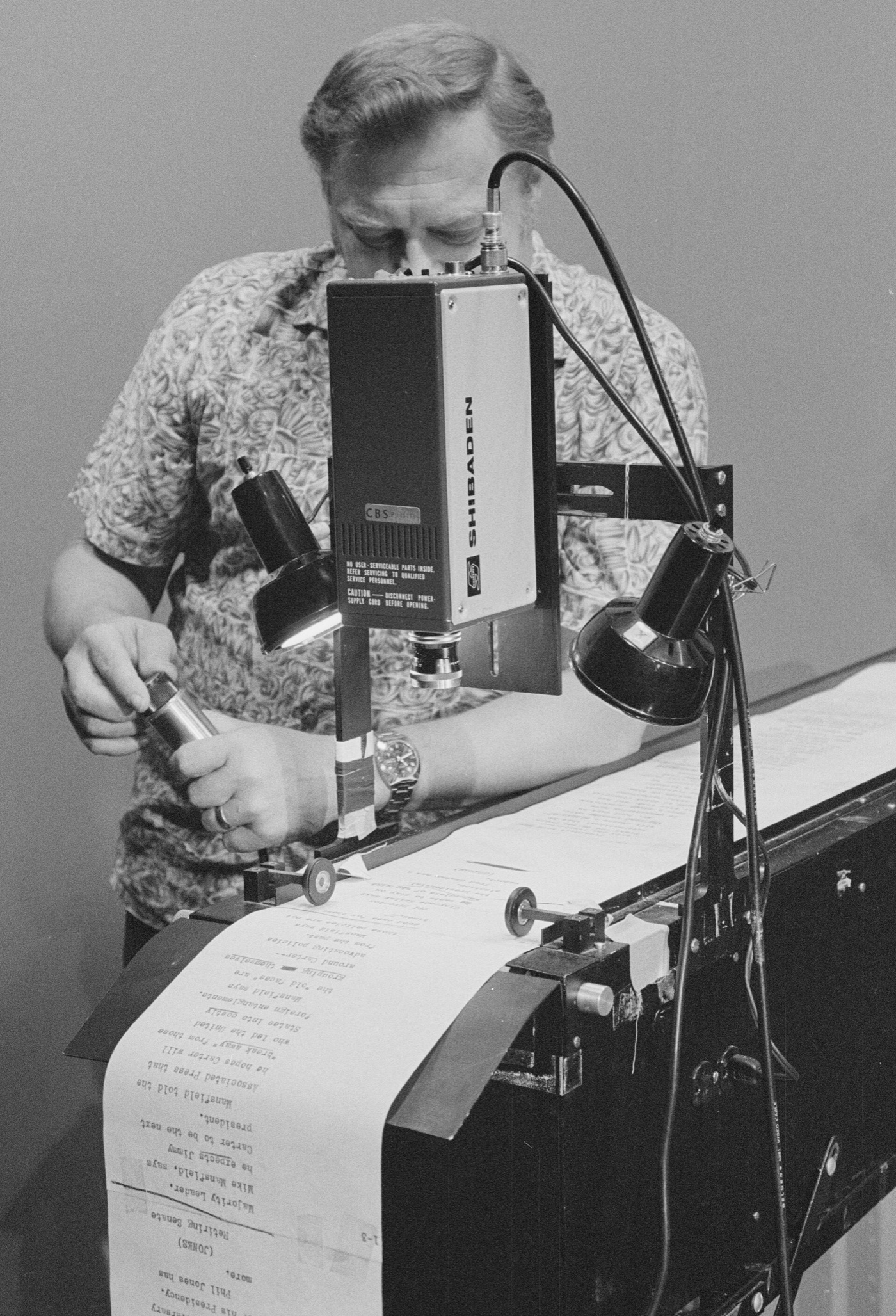 A man in a patterned shirt feeds a long paper roll of typed script into a camera-mounted teleprompter